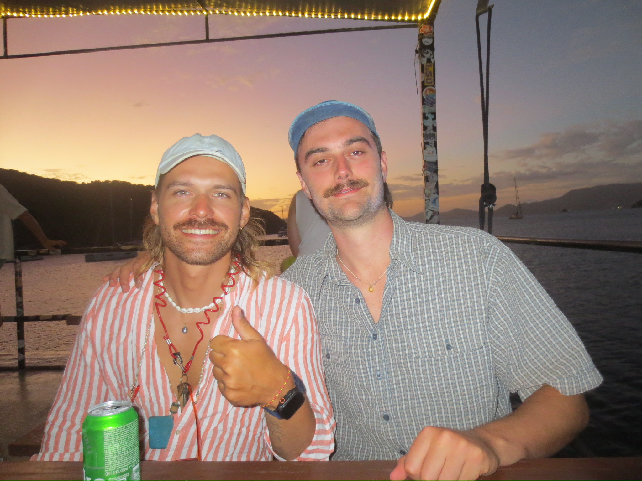 Two crew members smiling at a beach bar at sunset, one giving a thumbs up